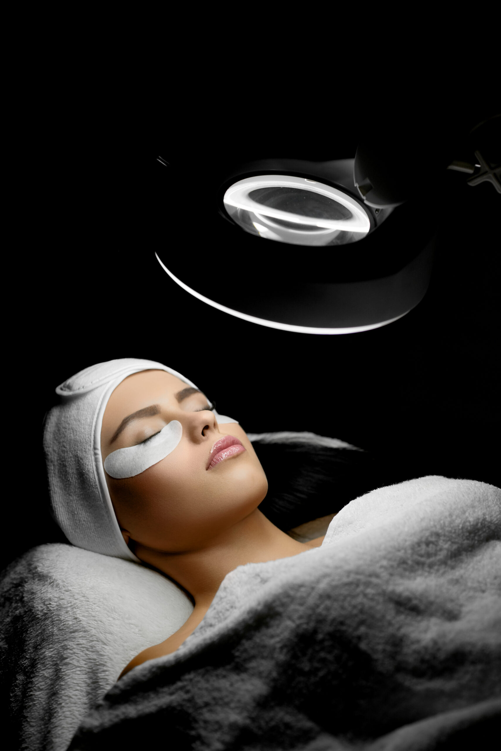 Woman lying on cosmetologiacal coach during lashes enlarging. Beaty salon procedure. young woman with pretty face wearing headbandage and stickers for lashmaking . Shadows in cosmetological cabinet.